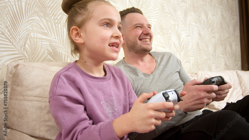 A father spends time with his little daughter, sitting on the sofa and playing a game console. Weekend activities, free time, home entertainment and video games concept. A father spends time with his little daughter, sitting on the sofa and playing a game console. Weekend activities, free time, home entertainment and video games concept.