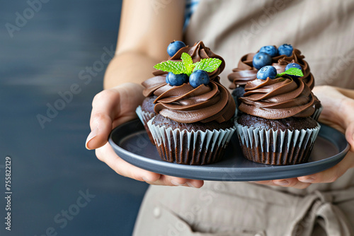 Woman in beige apron holding plate with chocolate cupcakes with brown whipped cream and decorated blueberry in confectionery. Copy space.