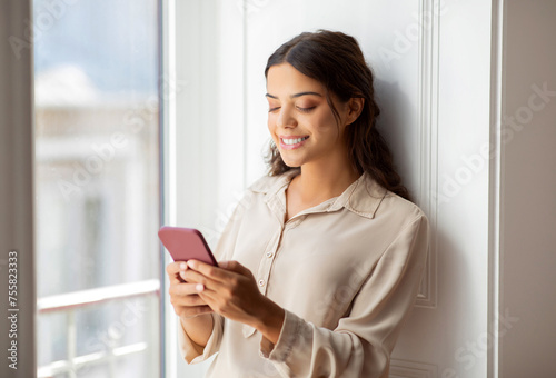 Beautiful Smiling Woman Standing By The Window And Using Smartphone