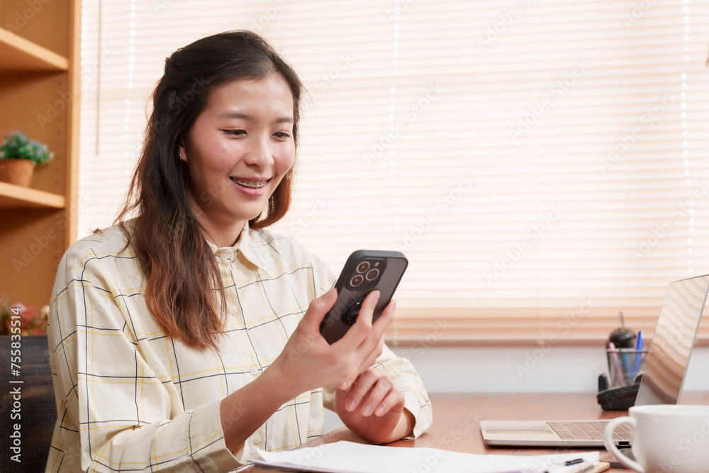 Joyful Asian lady beams at smartphone, experiencing pleasant chat in a work environment with a laptop and coffee on the table. Delighted by the content on her mobile device, exhibits a radiant smile Joyful Asian lady beams at smartphone, experiencing pleasant chat in a work environment with a laptop and coffee on the table. Delighted by the content on her mobile device, exhibits a radiant smile