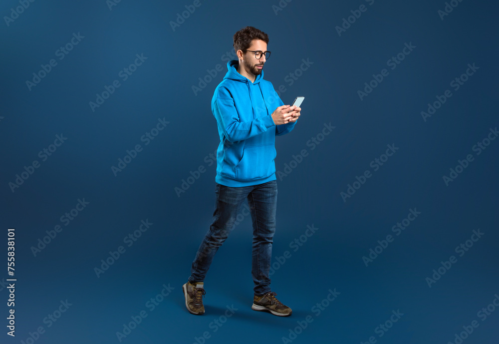 Young Stylish Man Walking With Smartphone In Hands Over Blue Studio Background - obrazy, fototapety, plakaty 