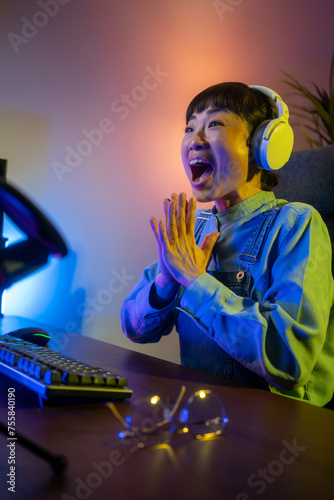 Excited Asian Gamer Girl in Headset with a Mic Playing Online Video Game on Her Personal Computer. She Talks to Other Players. Room and PC have Colorful Warm Neon Led Lights. Cozy Evening at Home.