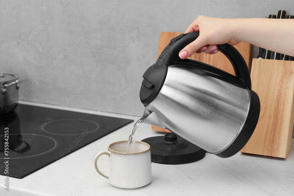Woman pouring hot water from electric kettle into cup in kitchen, closeup