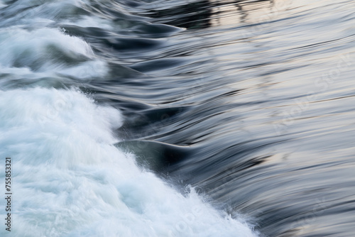 Rapid water flow over barrier with silky surface, river overflow over dam close up with splash and foam, abstract landscape pastel blue