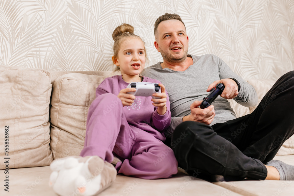 A father spends time with his little daughter, sitting on the sofa and playing a game console. Weekend activities, free time, home entertainment and video games concept. A father spends time with his little daughter, sitting on the sofa and playing a game console. Weekend activities, free time, home entertainment and video games concept.