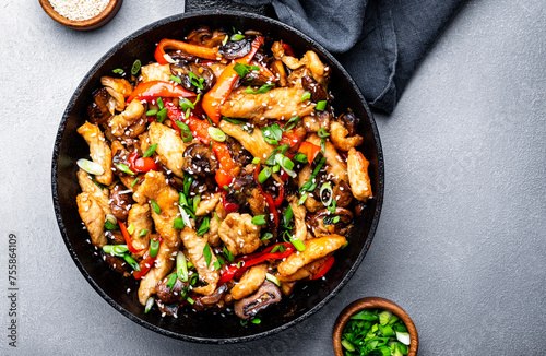 Asian stir fry chicken slices with red paprika, mushrooms, chives and sesame seeds in frying pan. Gray kitchen table background, top view