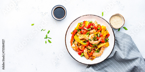 Spicy stir fry shrimps with colorful paprika, green peas, onion and sesame seeds with ginger, garlic and soy sauce in white bowl on kitchen table background, top view banner