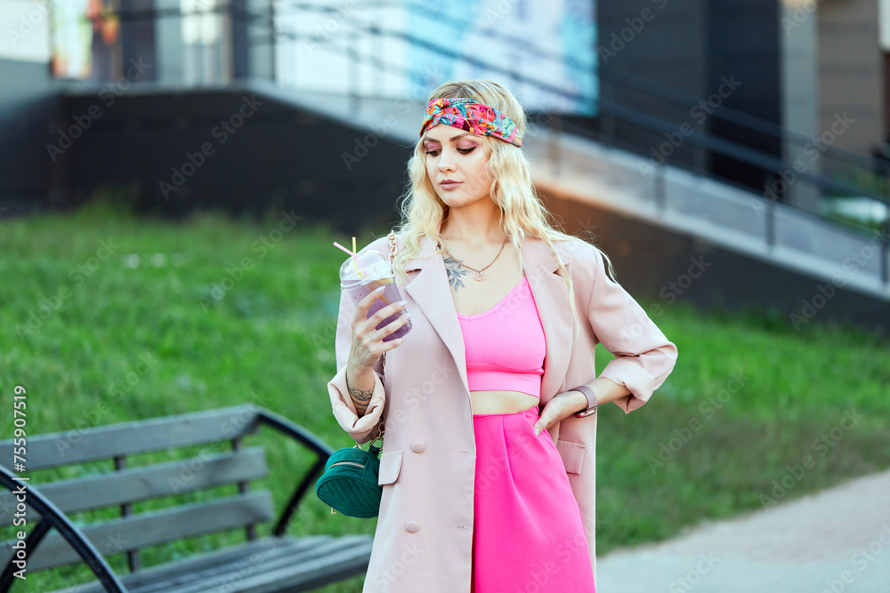 Girl holding a drink in her hands. Summer photo.Stylish female hipster walking on a street. Elegant woman wearing trendy pink suit and trousers, sunglasses, with textured green shoulder bag.