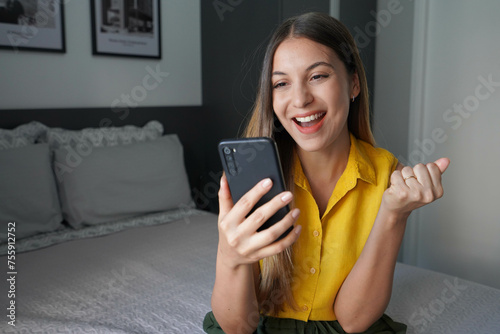 Happy young Brazilian woman holding smartphone and celebrates with a clenched fist sitting on bed at home