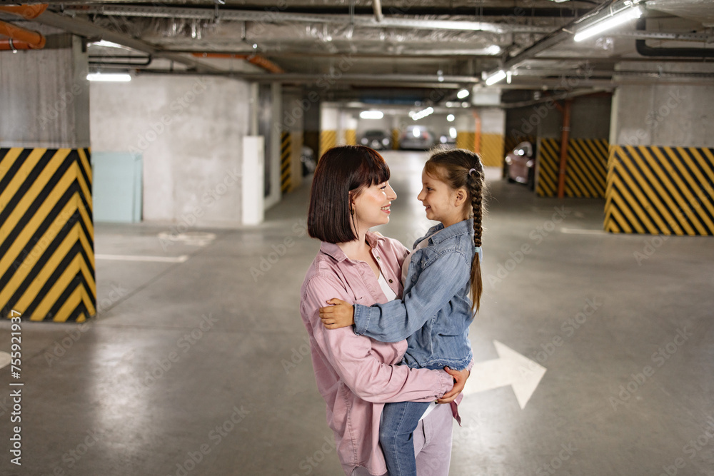 Portrait of Caucasian mother holding her preschool daughter in hands having fun in underground parking lot. Happy family in casual clothes walking to their car before an exciting trip.