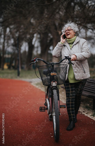 Cheerful mature lady with curly hair chatting on her smart phone while standing next to her bike in a park setting.