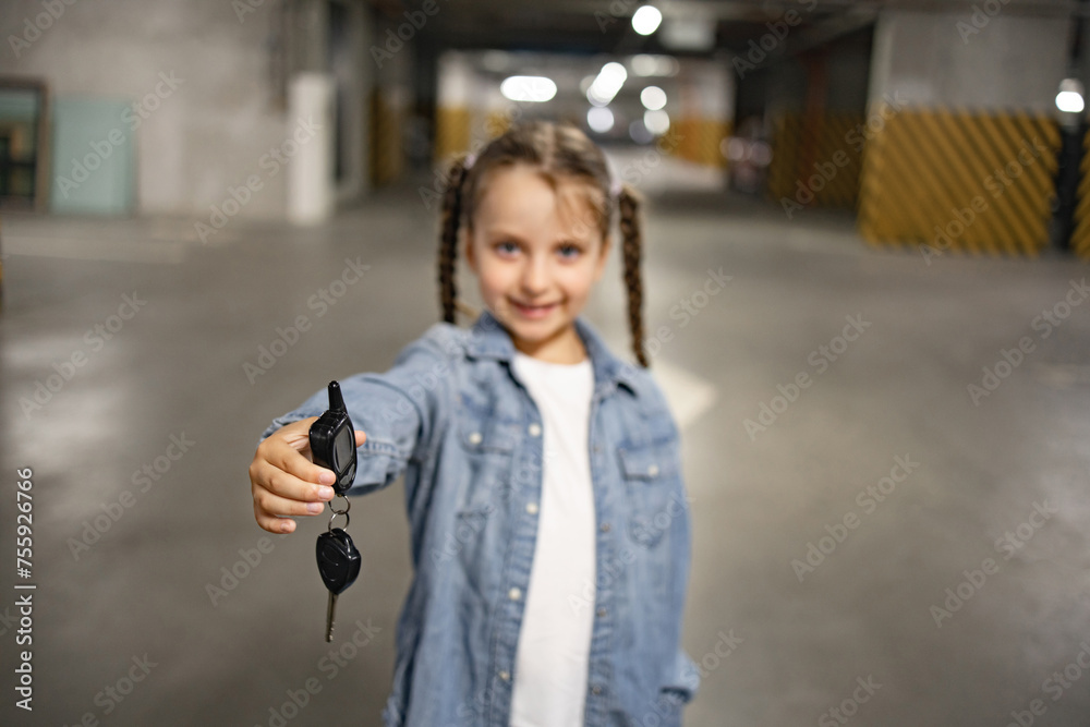 Selective focus of hand of cute little girl standing in underground parking lot in casual clothes holding keys opening car remotely, looking at camera.