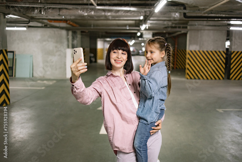 Caucasian mother holding preschool daughter in hands making selfie with smartphone in underground parking lot. Happy family in casual clothes having video call using cell phone before exciting trip.