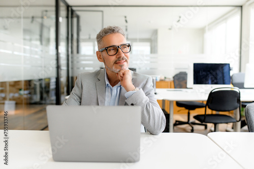 A contemplative businessman gazes into the distance from his laptop, portraying strategic thought in a serene, modern office setting, the blend of technology and human insight