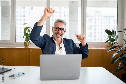 Excited mature businessman is gesturing victory with his arms raising up, happy senior ceo screaming with triumph, winning in game, having received great news of a good deal