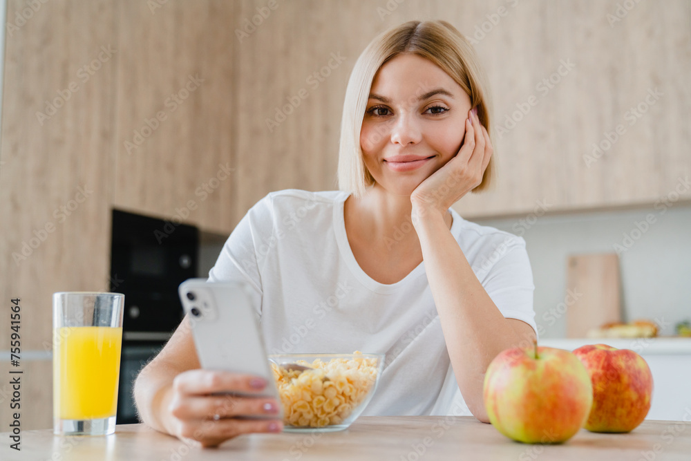 Young woman looking at the camera using cellphone, scrolling social media, doing online shopping while having breakfast. Caucasian girl eating cereals drinking orange juice Young woman looking at the camera using cellphone, scrolling social media, doing online shopping while having breakfast. Caucasian girl eating cereals drinking orange juice