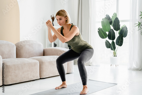 Caucasian female athlete doing squats on fitness mat at home. Strong fit woman on training, body slimming shaping, burning calories in the living room