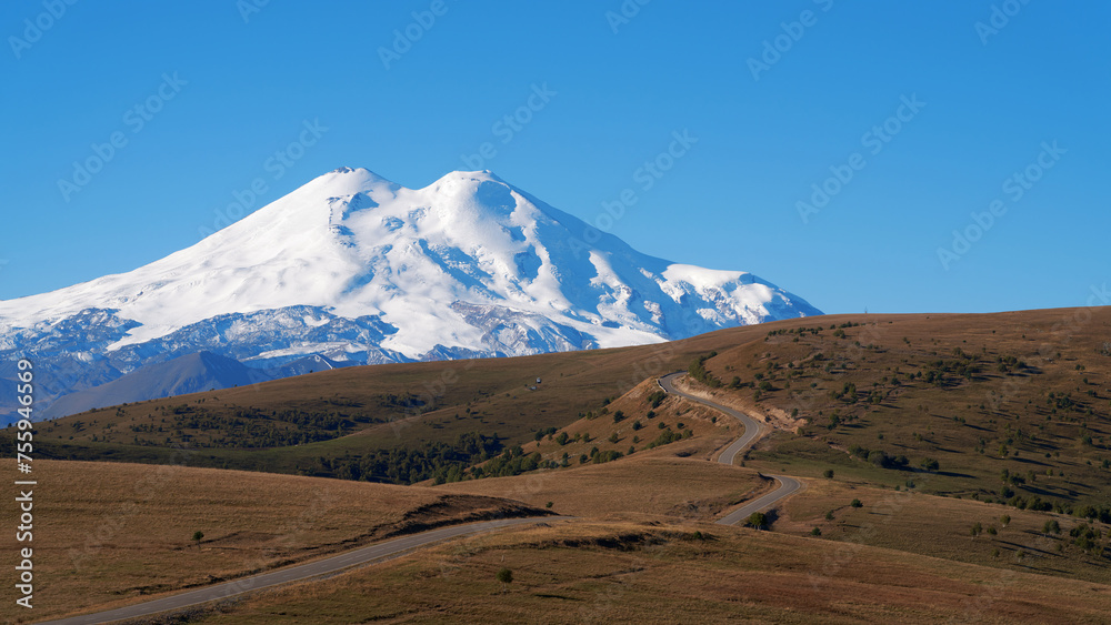 A narrow winding highway along the mountain plateau to the snowy Elbrus against the blue sky. Copy space.