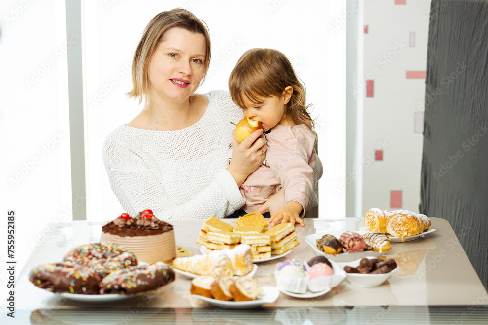 Woman giving apple to a child for snack, sitting beside table full of sweet food and unhealthy pastry with chocolate. Concept of parents choice wtah to give her child to eat, healthy lifestyle