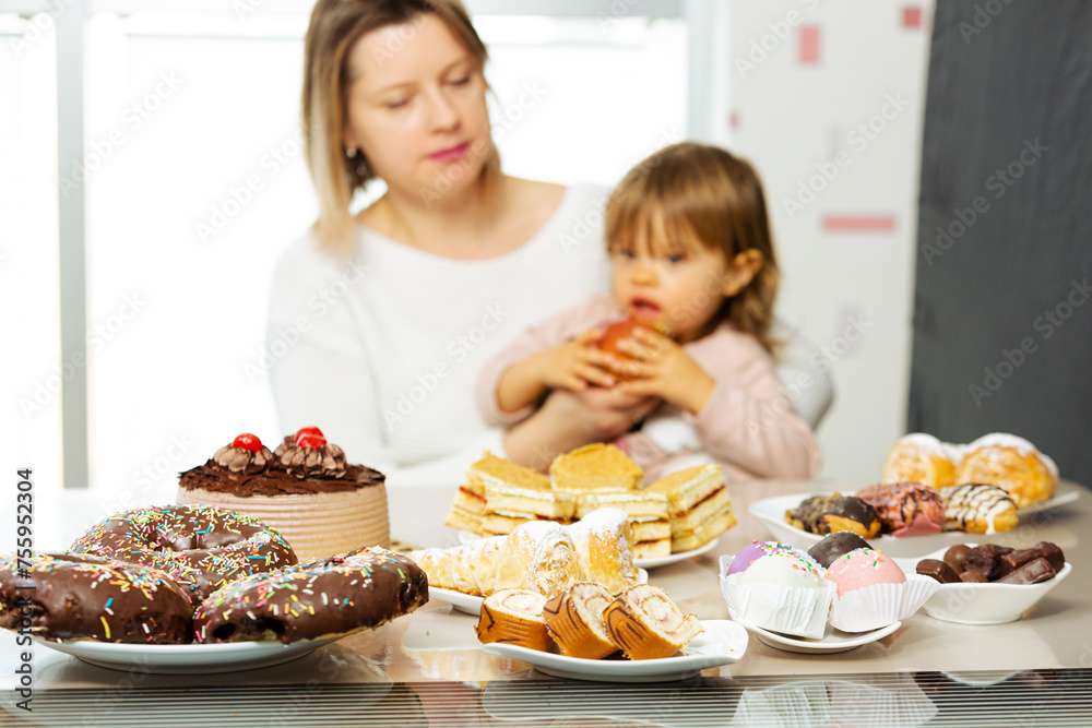 Woman giving apple to a child for snack, sitting beside table full of sweet food and unhealthy pastry with chocolate. Concept of parents choice wtah to give her child to eat, healthy lifestyle