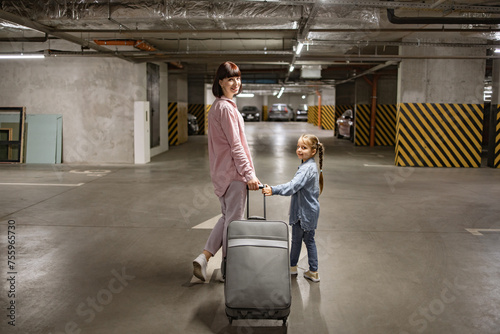 Youthful lady with her child clutching travel suitcase searching for vehicle before starting journey. Happy mom and small daughter, attired in informal wear, stroll through subterranean parking area. photo