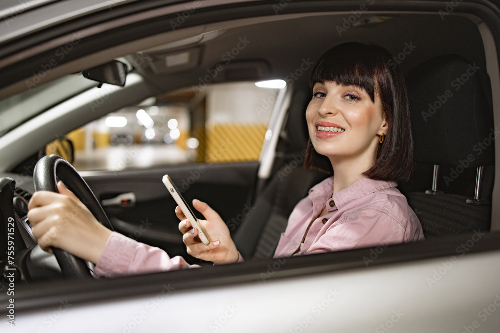 Attractive female typing on phone inside gray vehicle on underground parking lot. Young confident Caucasian woman in casual clothes sitting behind wheel of modern car with smartphone. Attractive female typing on phone inside gray vehicle on underground parking lot. Young confident Caucasian woman in casual clothes sitting behind wheel of modern car with smartphone.