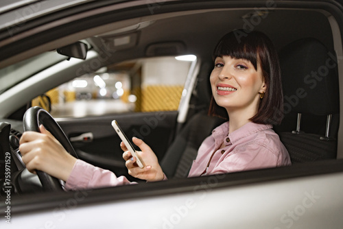 Attractive female typing on phone inside gray vehicle on underground parking lot. Young confident Caucasian woman in casual clothes sitting behind wheel of modern car with smartphone.