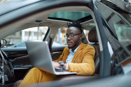 A young businessman sits on a car seat with a laptop.