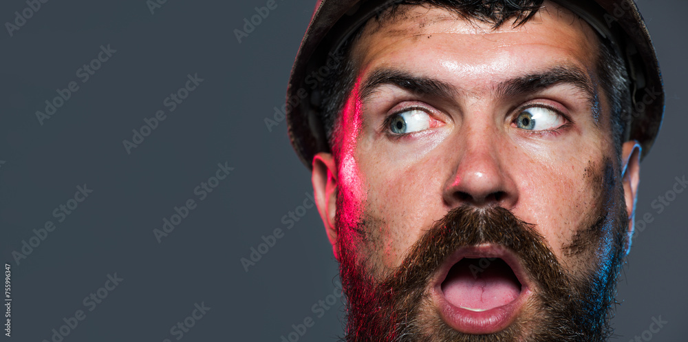 Closeup portrait of surprised construction worker in hard hat. Astonished bearded man in protective helmet. Amazed architect, contractor or male builder in safety hardhat. Copy space for advertising.