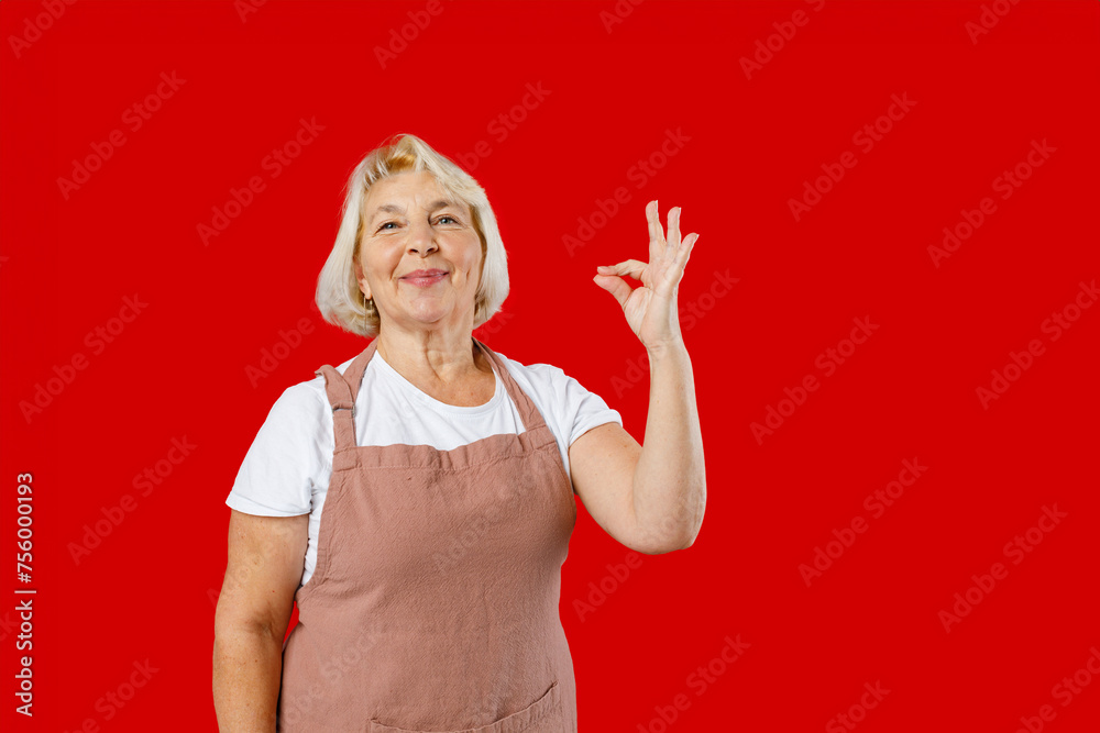 Elderly chef cooker housekeeper housewife woman in apron show sign of good taste, Bellissimo gesture, kiss fingers isolated plain on red color background. Elderly chef cooker housekeeper housewife woman in apron show sign of good taste, Bellissimo gesture, kiss fingers isolated plain on red color background.