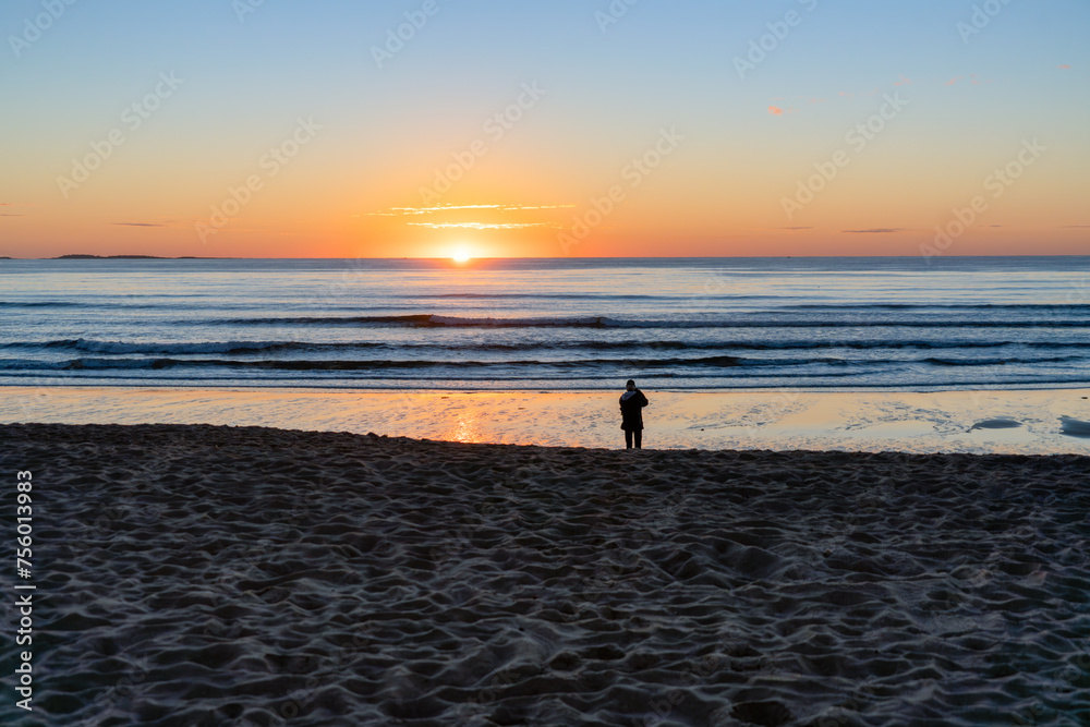 Silhouette of a person looking at and photographing a beautiful sunrise on the Old Orchard Beach in Maine.