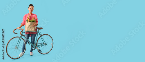 Young Asian woman with bicycle and bag full of fresh products on blue background with space for text