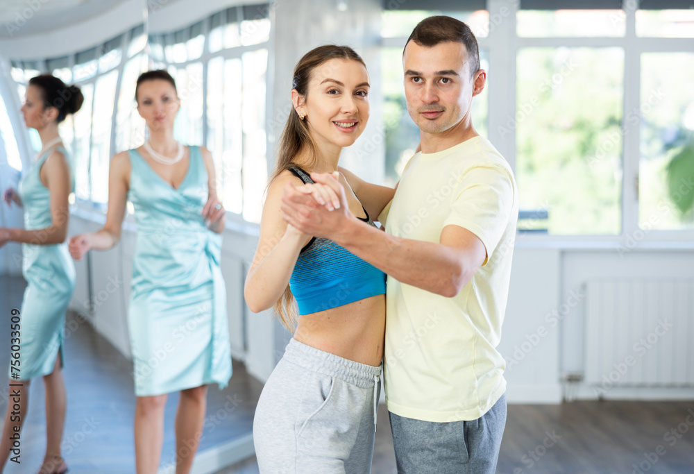 Smiling young adult couple in sportswear, bride and groom, rehearsing elegant wedding waltz in studio under guidance of professional female choreographer in bright studio..