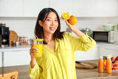 Beautiful Asian woman with glass of juice and oranges in kitchen