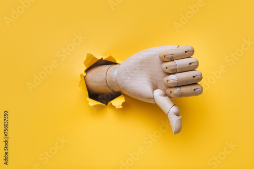 A wooden hand protrudes from a torn hole in yellow paper and shows a thumbs down (dislike). The concept of disapproval and that things will go badly. Artificial intelligence, robot.