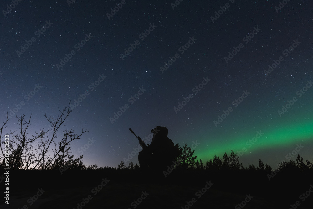 A special forces soldier with a night vision device and a rifle with a silencer is sitting in the forest against the background of the starry sky and northern lights.