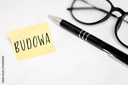 A yellow sticky note on a white background with the handwritten inscription "Budowa", next to it a black pen and glasses (selective focus) translation: construction