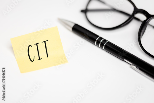 A yellow sticky note on a white background with the handwritten inscription "CIT", next to it a black pen and glasses (selective focus) 
