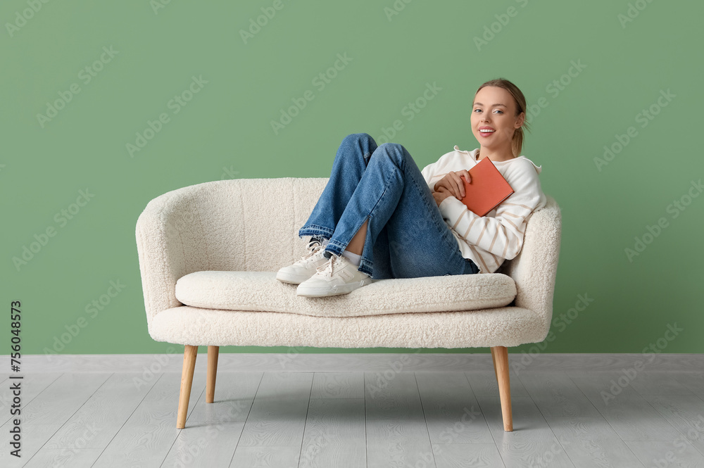 Beautiful young happy woman reading book on comfortable sofa near green wall