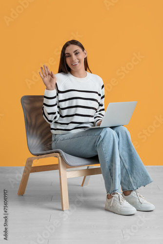 Beautiful young happy woman with laptop sitting on comfortable chair and waving hand near yellow wall