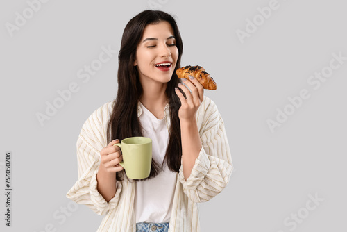 Beautiful young woman with cup of tea and croissant on grey background