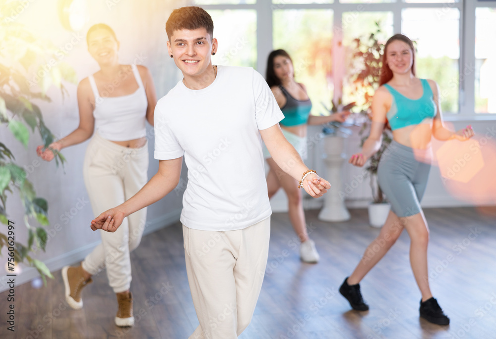 Fototapeta premium Positive smiling male teenager learns social dance groove, perform movements in choreographic class in company of peers. Young girls and guy repeat movements, train in spacious training hall.