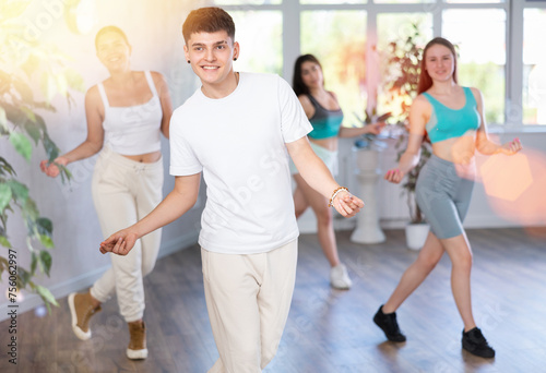 Positive smiling male teenager learns social dance groove, perform movements in choreographic class in company of peers. Young girls and guy repeat movements, train in spacious training hall.