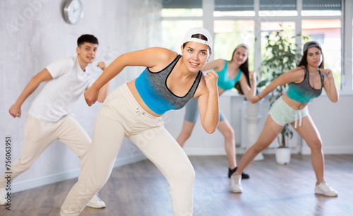 Female teenager in ball cap engaged shake leg in fitness studio with friends, young people in good mood actively move and dance hip hop at choreography lesson