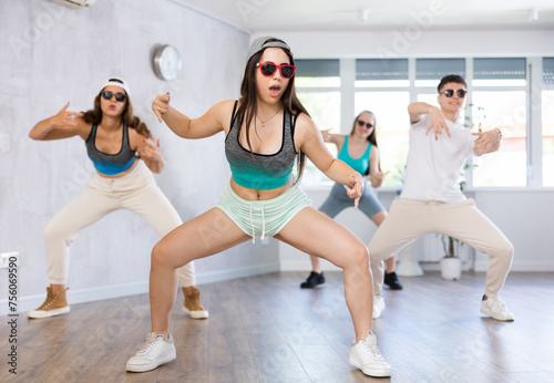 Positive female teenager dances jazz-funk in choreographic school, group of young people in sportswear and dark glasses ball cap train together with classmates in gym before competitions, battle
