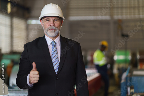 A man in a suit and a hard hat stands in front of a building. He is wearing a tie and a white helmet, show thumb up