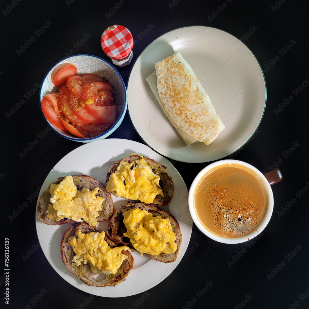The table top view presents a delightful home-cooked breakfast spread, featuring toasted bread with scrambled eggs, a peanut butter wrap, a cup of black coffee, and slices of fresh tomato. The table top view presents a delightful home-cooked breakfast spread, featuring toasted bread with scrambled eggs, a peanut butter wrap, a cup of black coffee, and slices of fresh tomato.