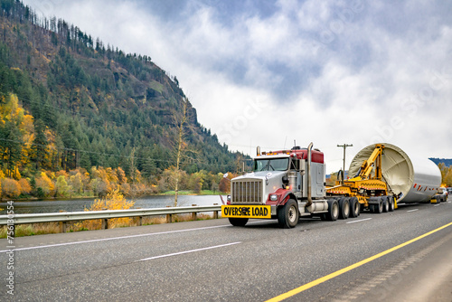 Big rig semi truck with oversize load sign on the bumper transporting oversized part of a wind turbine strut using an additional transport trolley running on the highway road along the Columbia River