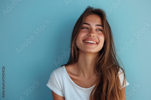 Cheerful young woman with long hair and white t-shirt smiling brightly against a blue background, exuding happiness and carefree youthfulness