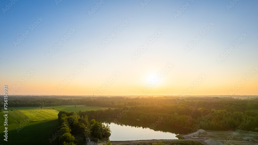 This photograph presents the first light of dawn breaking over a serene countryside landscape. A small lake sits quietly amidst fields and trees, reflecting the early morning sky. The soft light of